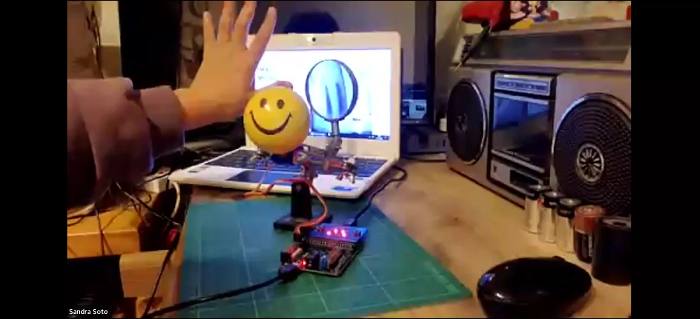 Sandra Soto raises her hand in front of a laptop camera, and a smiley-face creature mounted on a servo turns in response, wired to a micro:bit on her desk.